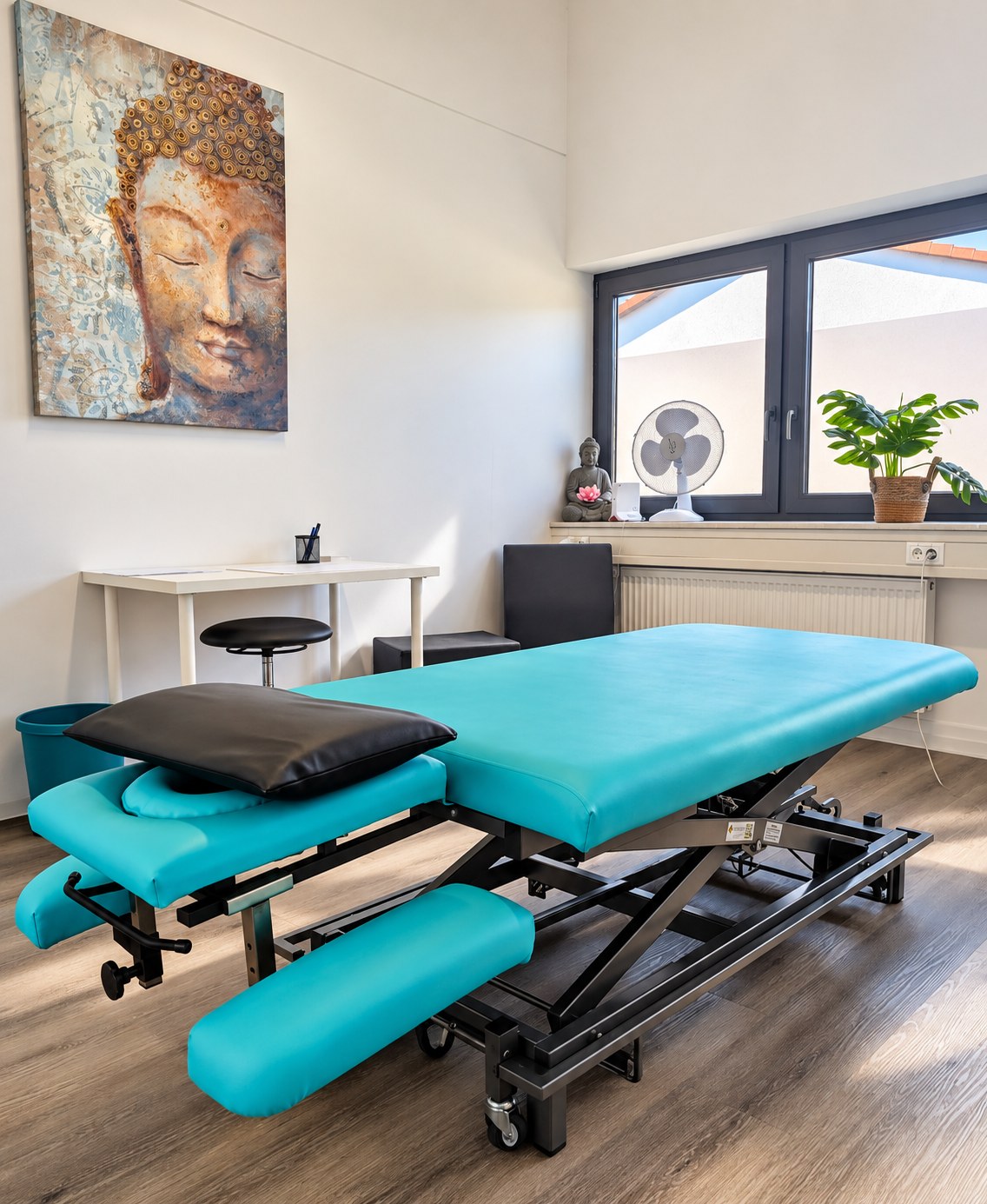 Heller Behandlungsraum mit türkisfarbener Therapieliege, Buddha-Wandbild und Fenster